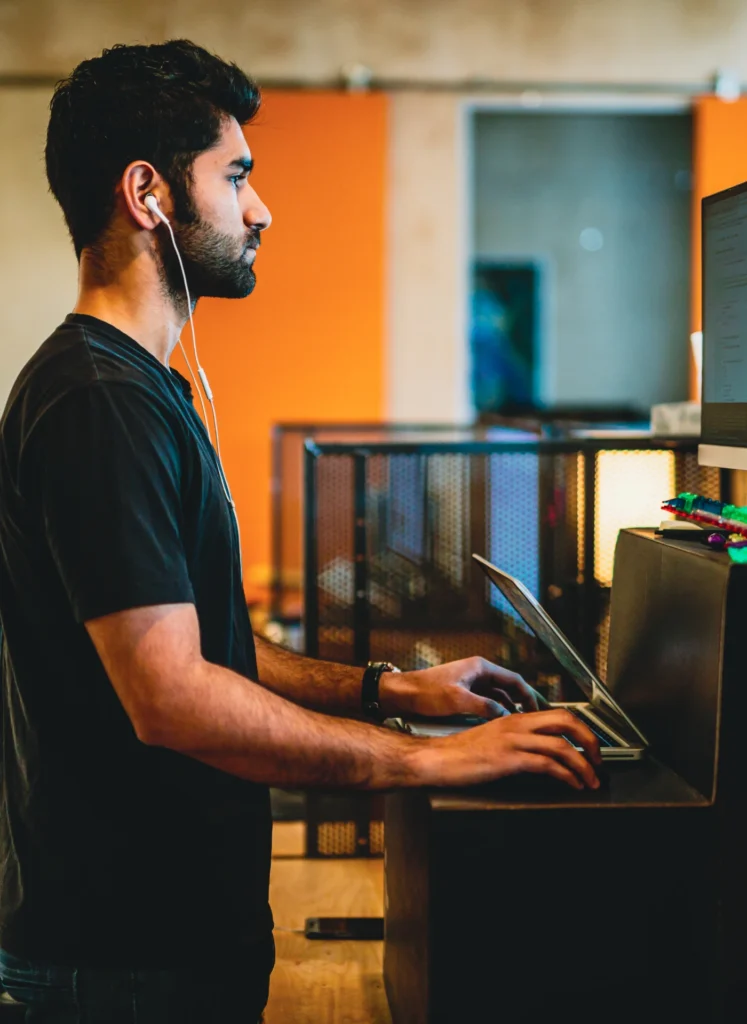 Man with white earphones typing on a laptop at a desk in a modern office, viewed from the side.