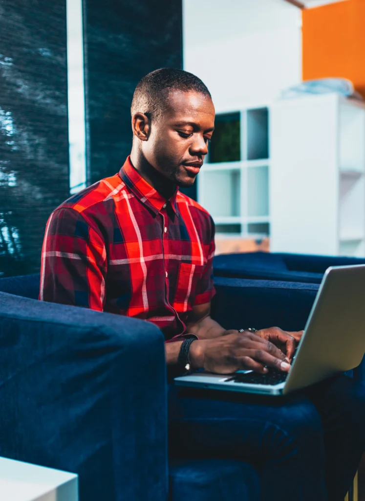 Man typing on a laptop while seated on a blue sofa in a red plaid shirt.