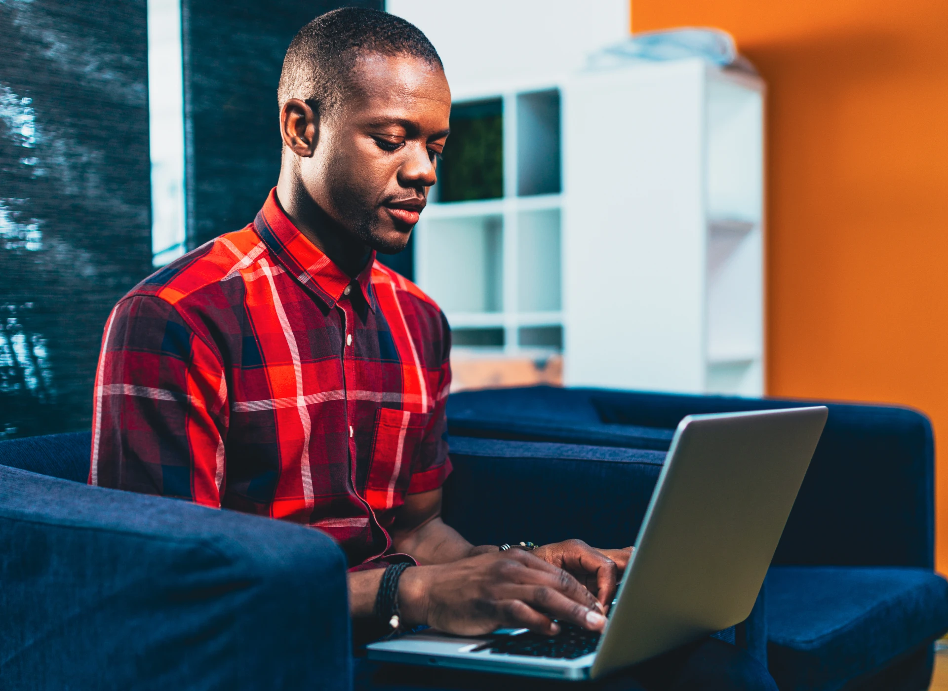 Man in a red plaid shirt typing on a laptop while sitting on a blue couch in a modern living room.