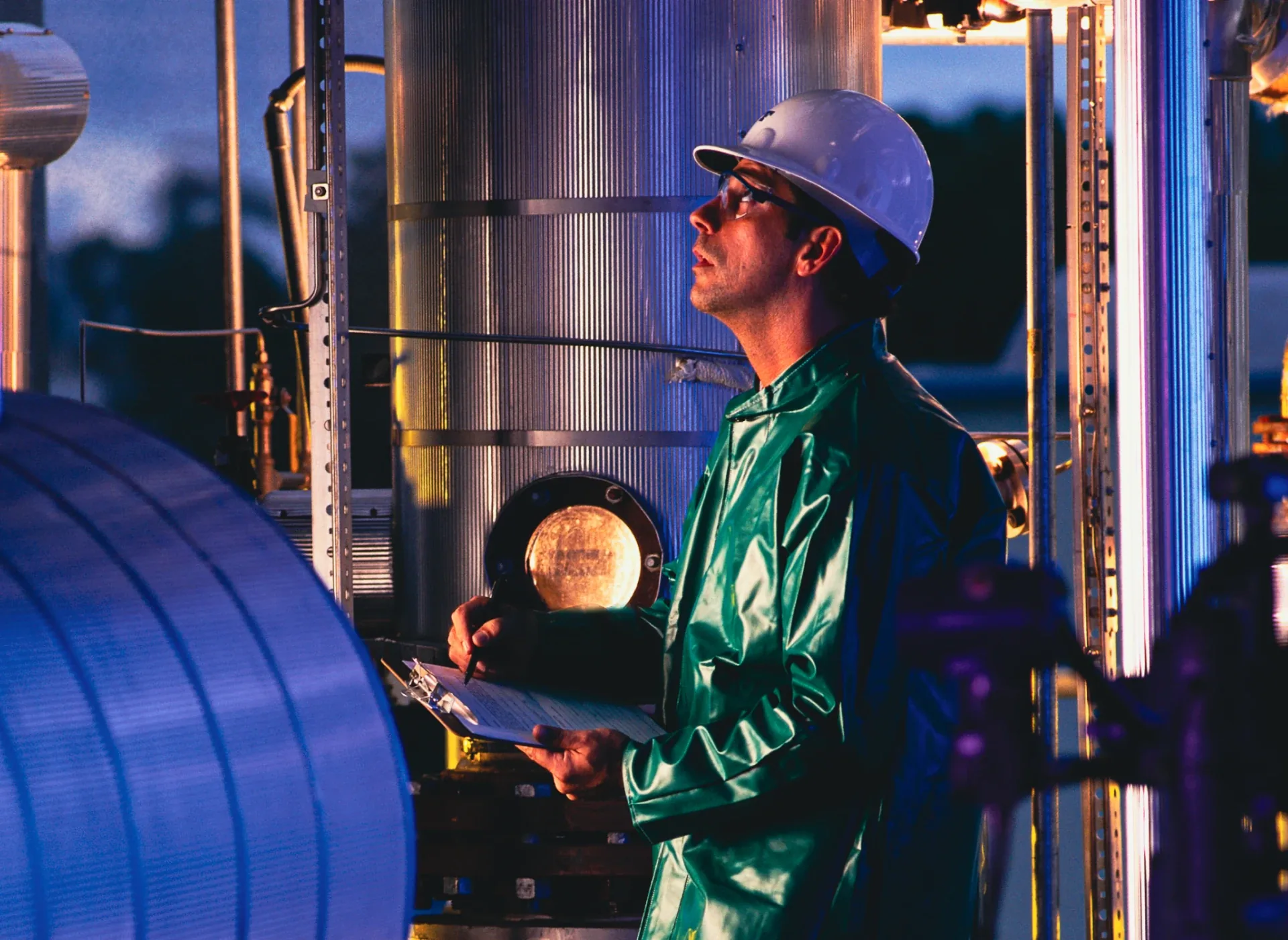Engineer in hard hat and green coveralls examines equipment, holding a clipboard in an industrial facility at dusk/dawn.