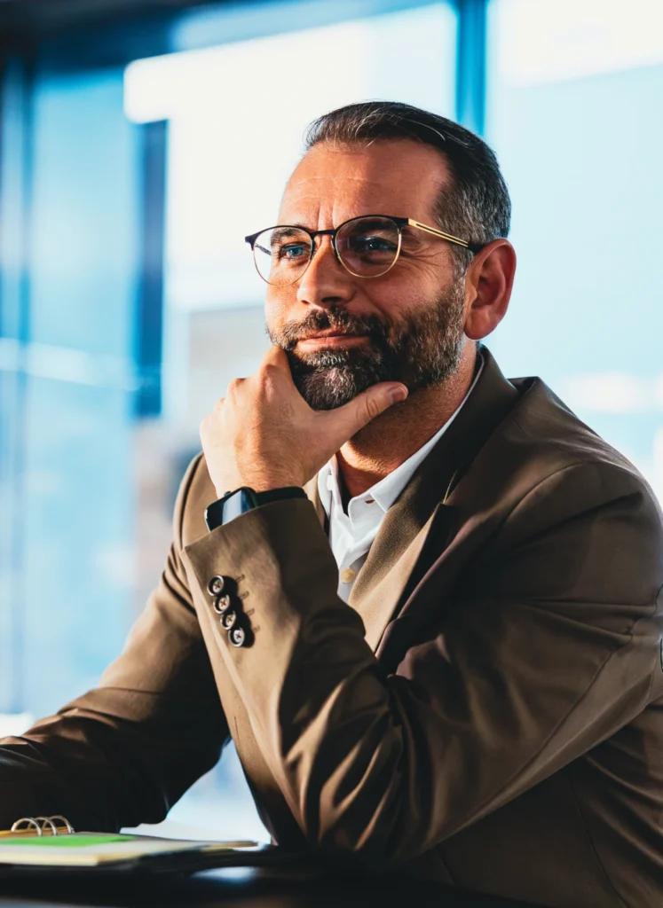 Man in a brown blazer and glasses, resting his chin on his hand in a thoughtful office setting.