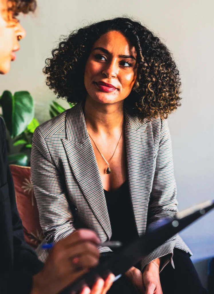 Woman in a checkered blazer smiling slightly, engaged in a conversation with a person in front of her.