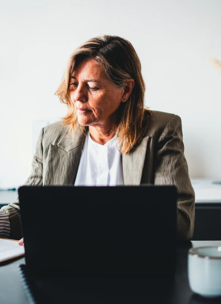 Woman in a blazer sits at a desk, focused on her laptop screen in an office setting.