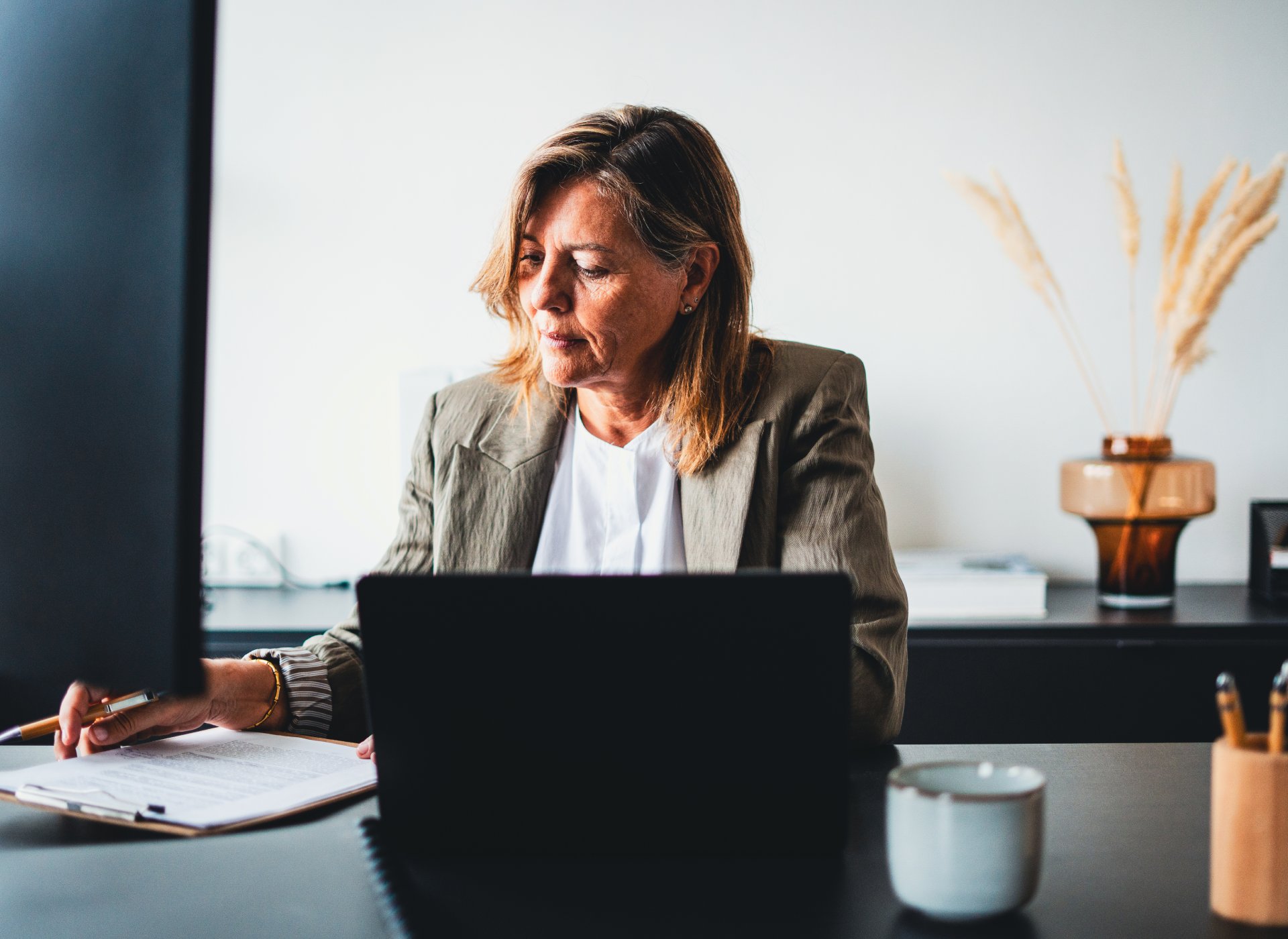Woman in a blazer sits at a desk, reviewing papers with a pen while a laptop sits in front of her in a bright office. A mug and decorative vase are on the desk behind her.