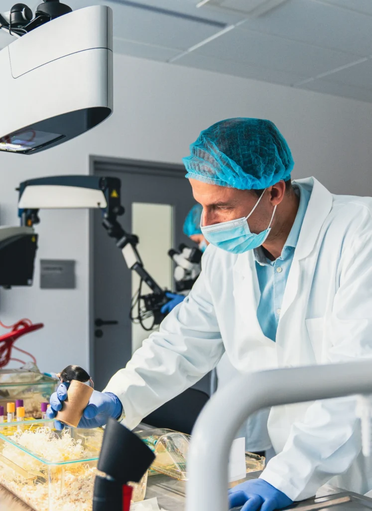 Lab technician in blue scrubs, mask, and hairnet working on a sample at a sterile lab bench under bright overhead light.