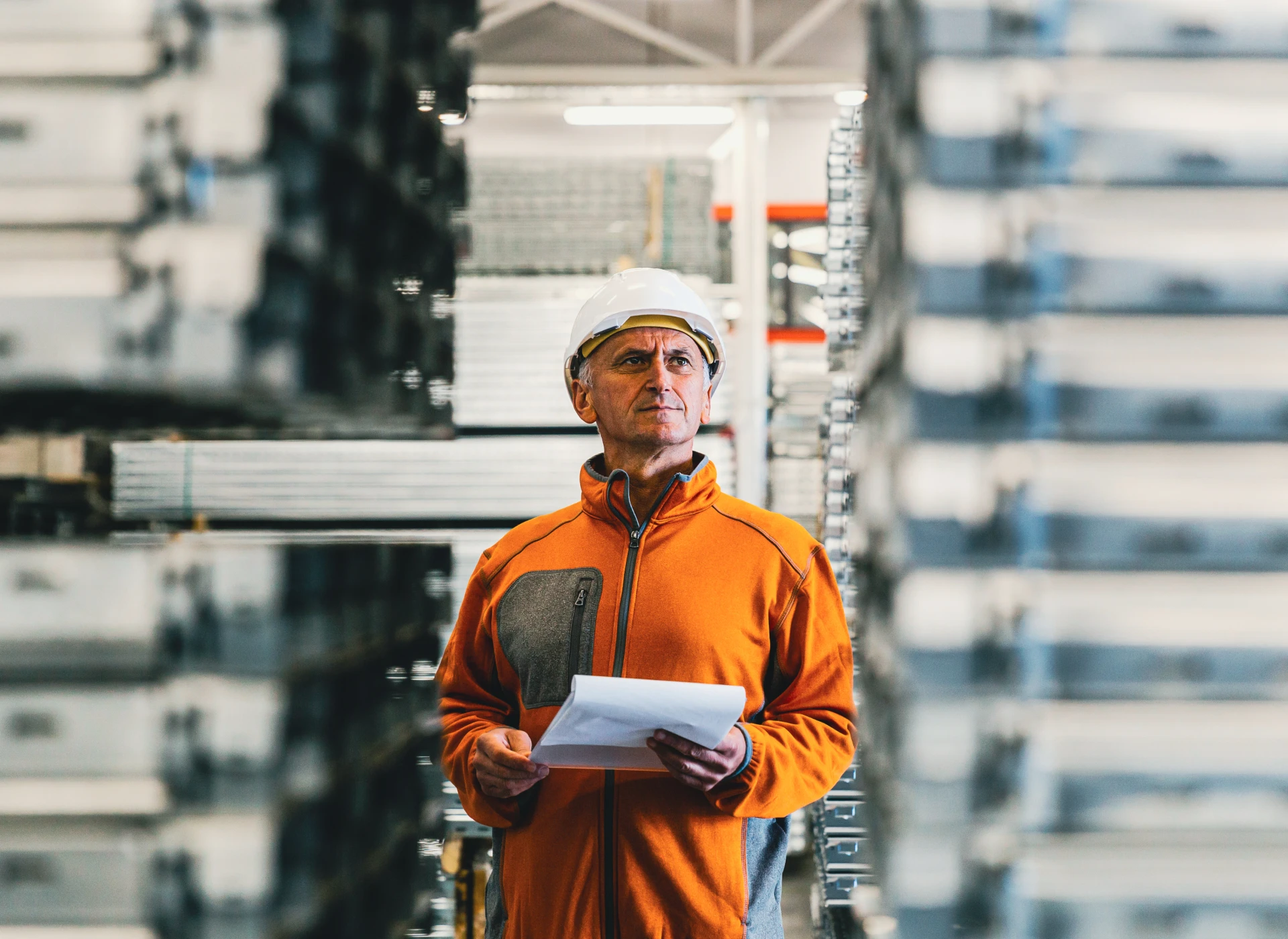 Warehouse worker in orange jacket and hard hat standing between shelves, holding papers, inspecting inventory.