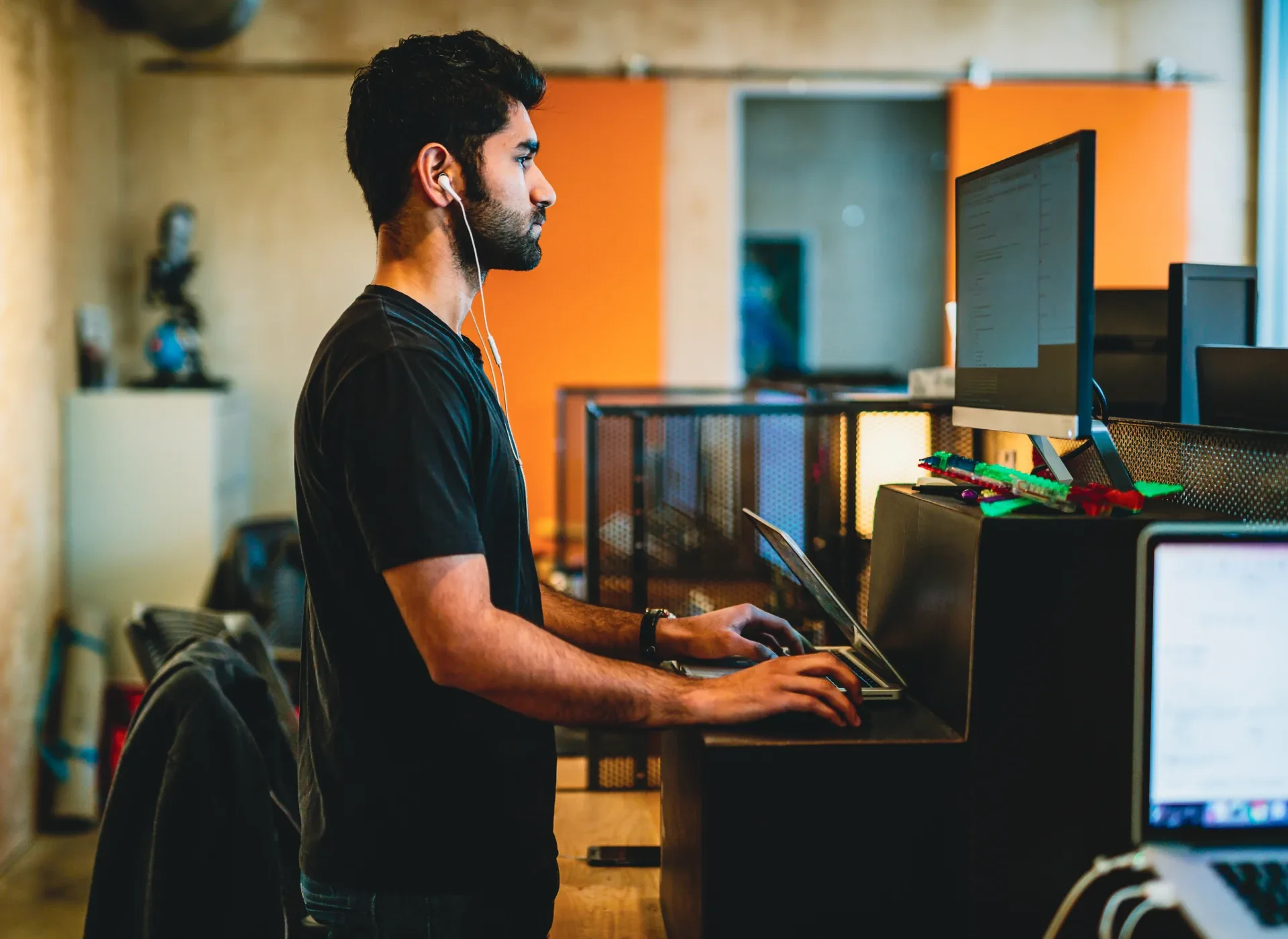 Man wearing white earbuds standing at a desk, typing on a laptop in a modern office setting.